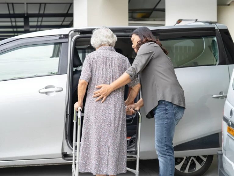Elderly woman with walker receiving assistance with transportation into a car.