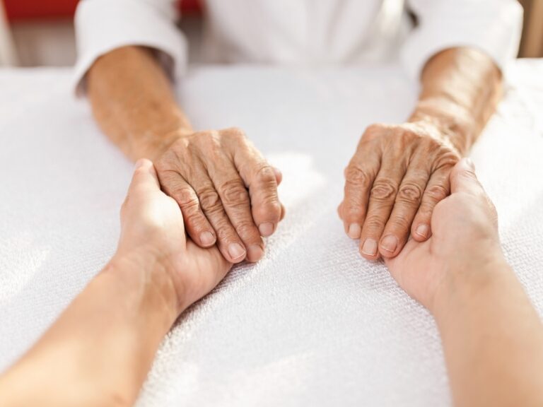 carer support dementia – caregiver holding elderly person’s hands on white table showing compassion and connection