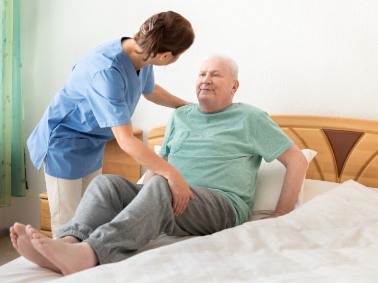Caregiver helping elderly man sit up in bed, showing attentive 24-hour personal care support.