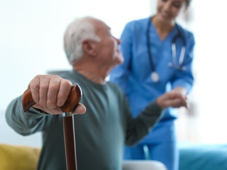 Elderly man receiving standard home care support from nurse in blue uniform holding his hand.