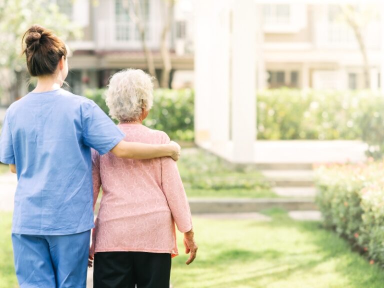 Caregiver walking with elderly woman in garden, symbolising compassion and home care comfort.